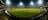 A brightly lit cricket stadium under the night sky with a lush green field, surrounded by multicolored seating and empty stands. A few people can be seen on the field, possibly players or staff.