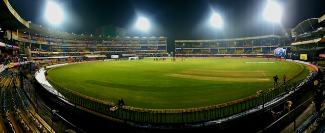 A brightly lit cricket stadium under the night sky with a lush green field, surrounded by multicolored seating and empty stands. A few people can be seen on the field, possibly players or staff.