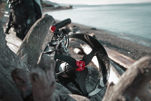 A dive scooter with a black and red finishing is positioned on driftwood at a rocky beach. The ocean stretches out in the background under a cloudy sky, with another piece of equipment partially visible to the left.