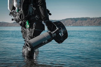 A scuba diver in a black suit stands waist-deep in water, holding an underwater scooter. The equipment includes a diving tank and other gadgets. The background features a calm body of water with a forested shoreline under a clear sky.