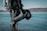 A scuba diver in a black suit stands waist-deep in water, holding an underwater scooter. The equipment includes a diving tank and other gadgets. The background features a calm body of water with a forested shoreline under a clear sky.