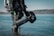 A scuba diver in a black suit stands waist-deep in water, holding an underwater scooter. The equipment includes a diving tank and other gadgets. The background features a calm body of water with a forested shoreline under a clear sky.