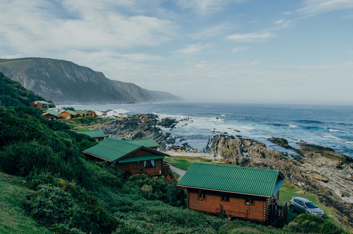 Green-roofed log cabins above a rocky shore in Tsitsikamma National Park, with a high headland behind