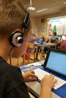 A classroom scene where a student seated at a desk is using a tablet with an external keyboard. The student is wearing headphones. In the background, other students are also using tablets and working at their desks. The classroom has educational posters on the walls and a whiteboard at the front.