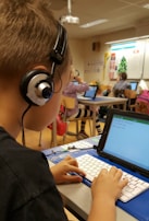 A classroom scene where a student seated at a desk is using a tablet with an external keyboard. The student is wearing headphones. In the background, other students are also using tablets and working at their desks. The classroom has educational posters on the walls and a whiteboard at the front.