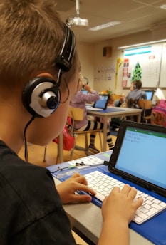 A classroom scene where a student seated at a desk is using a tablet with an external keyboard. The student is wearing headphones. In the background, other students are also using tablets and working at their desks. The classroom has educational posters on the walls and a whiteboard at the front.