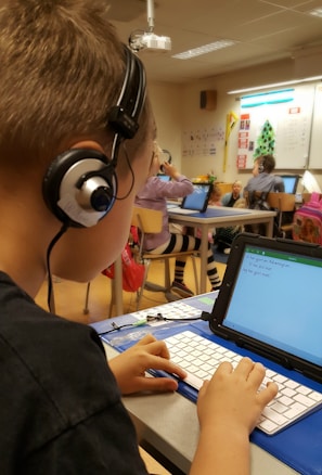 A classroom scene where a student seated at a desk is using a tablet with an external keyboard. The student is wearing headphones. In the background, other students are also using tablets and working at their desks. The classroom has educational posters on the walls and a whiteboard at the front.