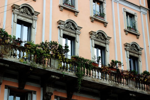 A classic European-style building with ornate architectural details, including decorative window frames and an elaborate balcony adorned with potted plants and flowering vines. The walls are painted in a soft peach color, adding warmth to the facade.