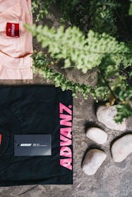 A black t-shirt with pink text 'ADVANZ' and a tag is placed on a textured stone surface alongside a group of smooth white stones. Above the t-shirt, a section of a peach-colored shirt with a red 'ADVANZ' label is visible. Green foliage is seen at the top and right edges, adding a natural element to the composition.