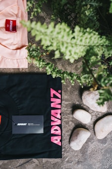 A black t-shirt with pink text 'ADVANZ' and a tag is placed on a textured stone surface alongside a group of smooth white stones. Above the t-shirt, a section of a peach-colored shirt with a red 'ADVANZ' label is visible. Green foliage is seen at the top and right edges, adding a natural element to the composition.
