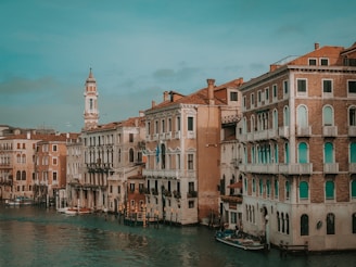 A picturesque view of a canal lined with historic Venetian buildings featuring ornate balconies and arched windows. A bell tower rises in the background, and the water is calm with reflections of the architecture.