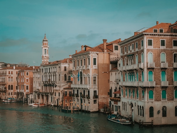 A picturesque view of a canal lined with historic Venetian buildings featuring ornate balconies and arched windows. A bell tower rises in the background, and the water is calm with reflections of the architecture.