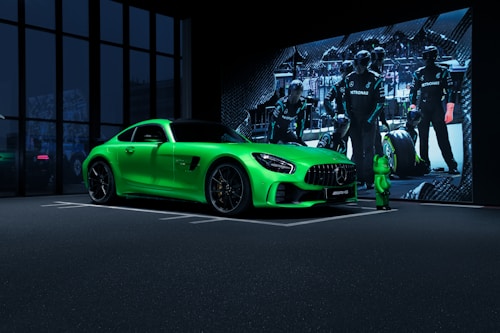 A vibrant green sports car is parked indoors near large glass windows. A projection on the wall behind features a pit crew wearing black uniforms with Mercedes branding, suggesting a motorsport theme.