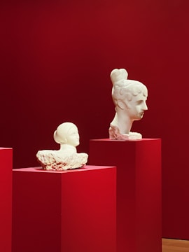 Two white marble busts are displayed on red pedestals against a deep red background. The larger bust features a classical hairstyle with detailed facial features, while the smaller one presents a simpler appearance with a headscarf.