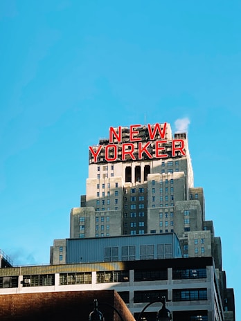 A large skyscraper with an art deco design features a prominent red sign that reads 'New Yorker' on its top. The building has multiple setbacks and a symmetrical façade with numerous windows. The sky is clear and blue, providing a vibrant backdrop.