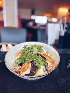 A bowl of fresh salad featuring green and red leafy lettuce, grilled chicken, walnuts, and sliced yellow fruit on a dark tabletop. The background displays a blurred indoor setting with warm lighting.