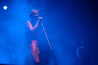 A person is singing into a microphone on stage, surrounded by a blue haze from the lighting and fog effects. The atmosphere is moody and ethereal, with the singer wearing a colorful outfit that stands out against the blue background. Another person can be faintly seen in the background.