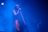 A person is singing into a microphone on stage, surrounded by a blue haze from the lighting and fog effects. The atmosphere is moody and ethereal, with the singer wearing a colorful outfit that stands out against the blue background. Another person can be faintly seen in the background.