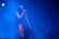 A person is singing into a microphone on stage, surrounded by a blue haze from the lighting and fog effects. The atmosphere is moody and ethereal, with the singer wearing a colorful outfit that stands out against the blue background. Another person can be faintly seen in the background.