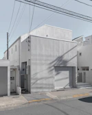 A modern, minimalist concrete building with a flat roof and small windows. The façade is smooth and featureless apart from some subtle texturing. In front, there is a gray garage door and a small entrance area. The structure is surrounded by other similar residential buildings, and power lines stretch across the sky above.