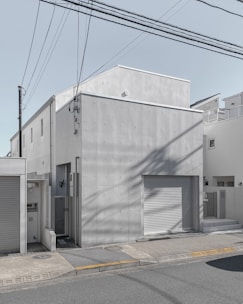 A modern, minimalist concrete building with a flat roof and small windows. The façade is smooth and featureless apart from some subtle texturing. In front, there is a gray garage door and a small entrance area. The structure is surrounded by other similar residential buildings, and power lines stretch across the sky above.