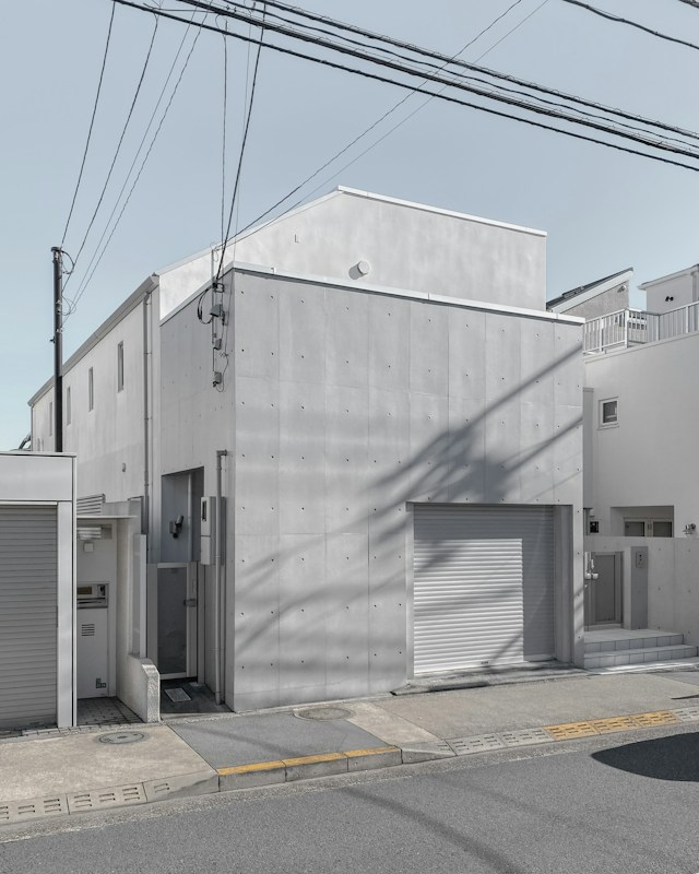 A modern, minimalist concrete building with a flat roof and small windows. The fa&ccedil;ade is smooth and featureless apart from some subtle texturing. In front, there is a gray garage door and a small entrance area. The structure is surrounded by other similar residential buildings, and power lines stretch across the sky above.
