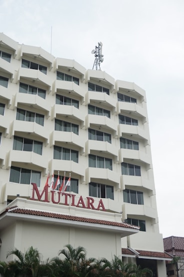 A multi-story building with a beige facade and numerous windows. There's a red sign with the word 'MUTIARA' above the entrance. The roof has a tiled design and the top of the building has antennas. Several small flags and palm trees are visible in front of the entrance.
