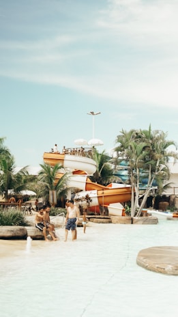 A water park scene with several people enjoying a warm day. There are slides in the background, surrounded by palm trees and other tropical plants. Some individuals are sitting at the edge of a shallow pool while others stand nearby. The sky is clear with some light clouds.