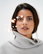 A close-up portrait of a woman with long dark hair, wearing a light gray knit sweater with a turtleneck. She holds a lit sparkler in front of her face, creating a playful and dynamic effect with the light reflections.