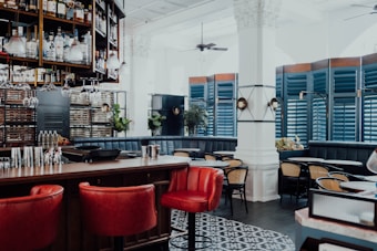 An upscale bar and lounge area with a modern aesthetic, featuring a well-stocked bar with glasses hanging overhead and a variety of bottles on display. The seating includes rich red leather bar stools and dark-colored cushioned booths, complemented by wooden tables and chairs with woven seats. The decor includes blue shutters, ornate columns, and hanging light fixtures, adding to the sophisticated ambiance.