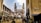 A busy street scene features a historic monument with four minarets. Surrounding the structure are shops and vendors, with numerous people walking and engaging in daily activities. The architecture and bustling atmosphere suggest a vibrant, culturally rich setting.