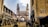 A busy street scene features a historic monument with four minarets. Surrounding the structure are shops and vendors, with numerous people walking and engaging in daily activities. The architecture and bustling atmosphere suggest a vibrant, culturally rich setting.
