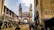 A busy street scene features a historic monument with four minarets. Surrounding the structure are shops and vendors, with numerous people walking and engaging in daily activities. The architecture and bustling atmosphere suggest a vibrant, culturally rich setting.
