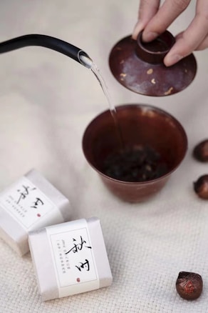 A hand lifts the lid of a small ceramic teapot as hot water is poured into it from a black-spouted kettle. Two neatly wrapped packets with Asian script are placed on a textured, cream-colored surface. Several round, brownish objects, likely tea balls or herbs, are scattered nearby.