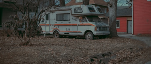 A vintage camper van is parked on a grassy area surrounded by bare trees and minimal foliage. The van has a weathered appearance with orange and brown stripes along its length. It is situated adjacent to a house with a red wooden exterior.