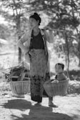 A woman is balancing a shoulder pole with baskets on each end, one carrying firewood and the other a young child. She stands on a dirt path shaded by tall trees, dressed in traditional clothing with a serene expression.