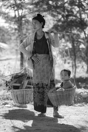 A woman is balancing a shoulder pole with baskets on each end, one carrying firewood and the other a young child. She stands on a dirt path shaded by tall trees, dressed in traditional clothing with a serene expression.