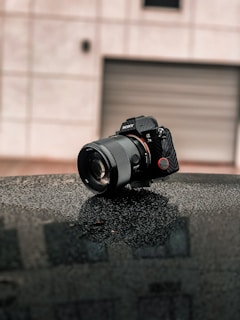 A digital camera with a large lens is positioned on a wet, dark surface, possibly a car roof. The background is blurred, showing an exterior wall with a closed garage door. The camera is angled, capturing the light reflections on its surface.
