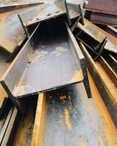 A pile of large, rusted metal I-beams stacked haphazardly outdoors. The beams show significant signs of oxidation, with orange rust contrasting against the dark gray of the metal. The surrounding area appears to be an industrial setting.