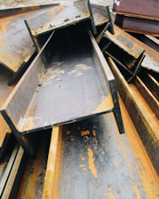 A pile of large, rusted metal I-beams stacked haphazardly outdoors. The beams show significant signs of oxidation, with orange rust contrasting against the dark gray of the metal. The surrounding area appears to be an industrial setting.