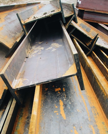 A pile of large, rusted metal I-beams stacked haphazardly outdoors. The beams show significant signs of oxidation, with orange rust contrasting against the dark gray of the metal. The surrounding area appears to be an industrial setting.