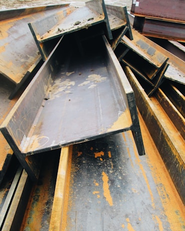 A pile of large, rusted metal I-beams stacked haphazardly outdoors. The beams show significant signs of oxidation, with orange rust contrasting against the dark gray of the metal. The surrounding area appears to be an industrial setting.
