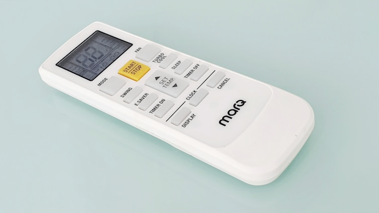 A white remote control with a digital display and several buttons, including a prominent yellow 'START/STOP' button, designed for operating an electronic device. The brand name 'marQ' is visible on the lower part of the remote. The buttons on the remote include functions for mode, swing, e-saver, timer, display, and others.