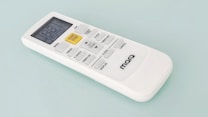 A white remote control with a digital display and several buttons, including a prominent yellow 'START/STOP' button, designed for operating an electronic device. The brand name 'marQ' is visible on the lower part of the remote. The buttons on the remote include functions for mode, swing, e-saver, timer, display, and others.