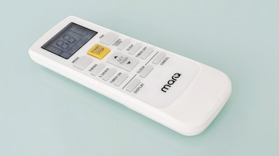 A white remote control with a digital display and several buttons, including a prominent yellow 'START/STOP' button, designed for operating an electronic device. The brand name 'marQ' is visible on the lower part of the remote. The buttons on the remote include functions for mode, swing, e-saver, timer, display, and others.