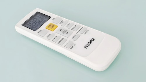 A white remote control with a digital display and several buttons, including a prominent yellow 'START/STOP' button, designed for operating an electronic device. The brand name 'marQ' is visible on the lower part of the remote. The buttons on the remote include functions for mode, swing, e-saver, timer, display, and others.