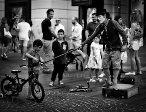 A street performer stands on a small platform, entertaining a crowd. He is wearing a hat and vest, extending a long stick towards a young boy with a bicycle. The onlookers, including other children and adults, mostly appear relaxed and intrigued as they watch the performance.