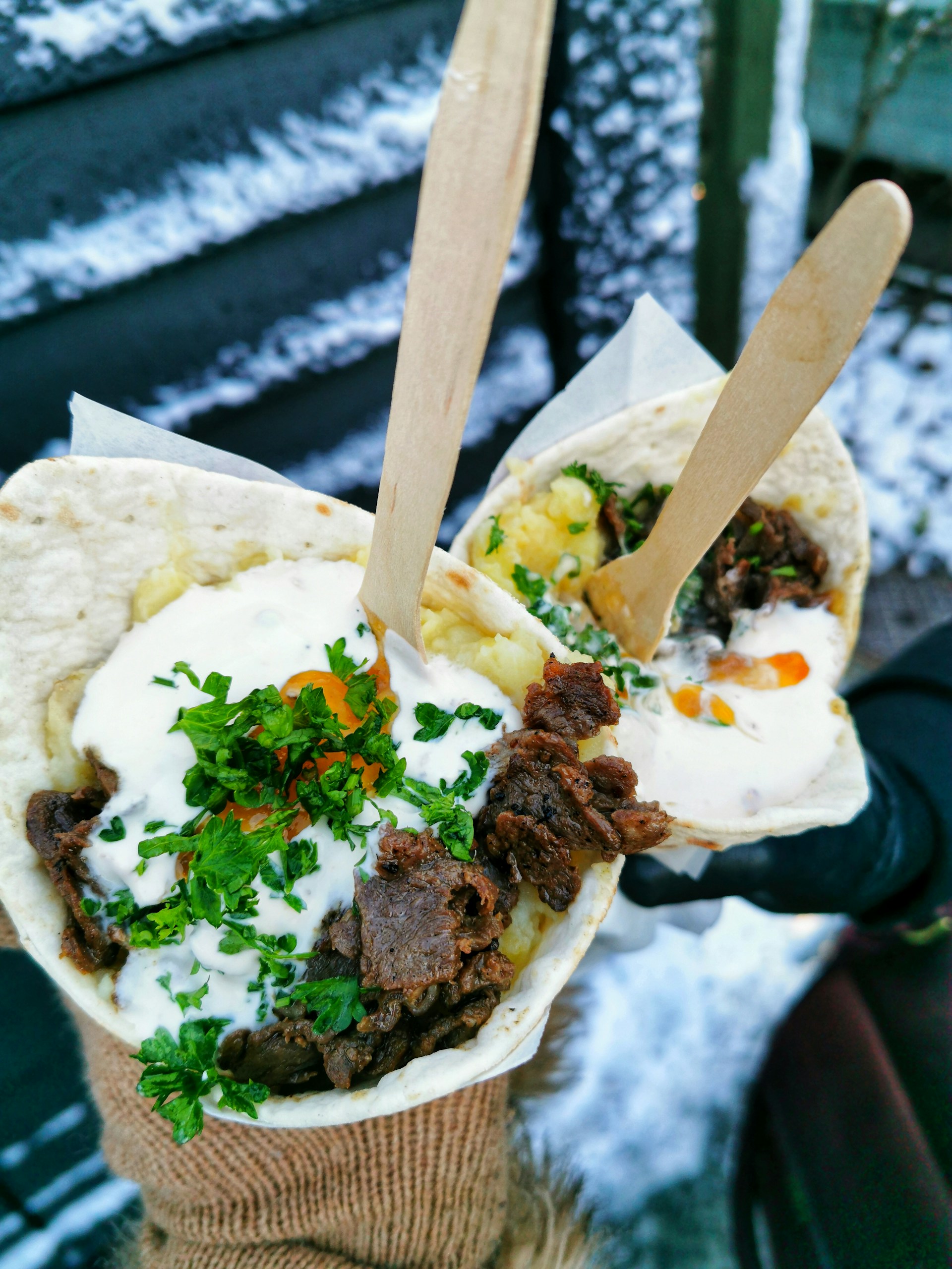 Two handheld wraps stuffed with grilled meat, fresh herbs, and creamy sauce are held outdoors. The background shows snow-covered surroundings, suggesting a winter setting. The wraps are garnished with parsley and sliced carrots, and are held using wooden forks. One hand is wearing a brown glove.