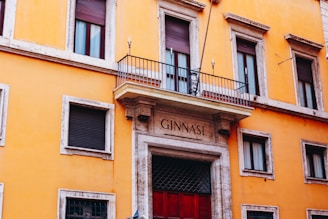 A historic yellow building with stone trim and multiple windows featuring closed shutters. A balcony with metal railing is centered above a large doorway, which is framed by stone columns. The words 'GINNASI' are engraved above the entrance, and a small decorative sign hangs in the center of the balcony.