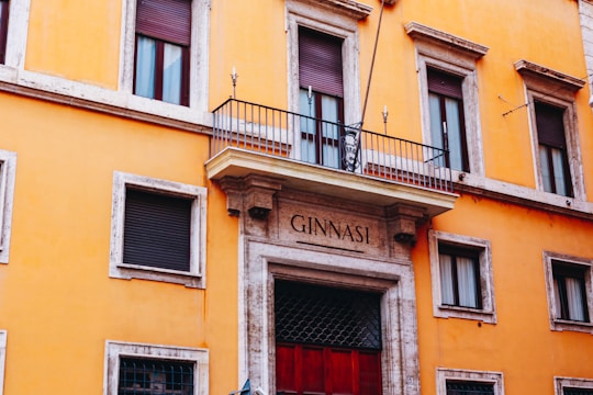 A historic yellow building with stone trim and multiple windows featuring closed shutters. A balcony with metal railing is centered above a large doorway, which is framed by stone columns. The words 'GINNASI' are engraved above the entrance, and a small decorative sign hangs in the center of the balcony.